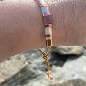 Könnte beinhalten: Armband mit rechteckigen Perlen in Rosa, Weiß und Roségold. Das verstellbare Band ist roségoldfarben, mit einer kleinen rosa Perle. Die Perlen sind etwa 0,5 cm lang. Mit Schiebeknotenverschluss.