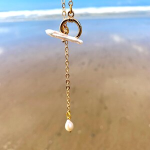 May include: A gold-toned chain necklace featuring a toggle clasp closure with a horizontal white stick-shaped pearl attached to the ring. A delicate gold chain extends downwards from the toggle, ending in a small, teardrop-shaped white pearl pendant.