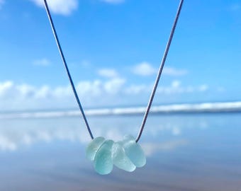 May include: A delicate necklace featuring a silver chain suspending a cluster of five pale blue sea glass pieces. The necklace is set against a backdrop of a bright blue sky and ocean waves.