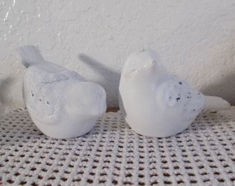May include: Two white ceramic bird figurines with a distressed finish. The birds are perched on a white crocheted surface.