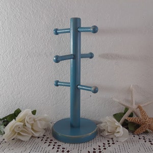 May include: A blue wooden mug tree with five hooks for hanging mugs. The mug tree is on a white lace tablecloth with white roses and starfish.
