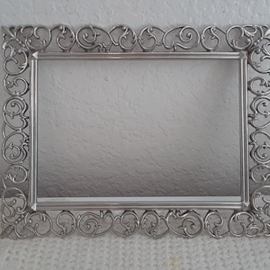 May include: A rectangular silver-colored picture frame with ornate scrollwork detailing. The frame has a classic design with decorative flourishes along the edges and corners. The frame is empty, ready to display a photograph or artwork.