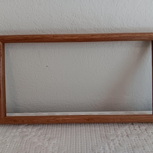 May include: A rectangular wooden picture frame with a medium brown finish and a visible natural wood grain texture. The frame features a simple, clean-lined design with mitered corners, providing a sturdy structure for displaying artwork or photographs.