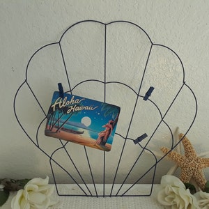 May include: A blue wire seashell shaped photo holder with a postcard that says "Aloha Hawaii" and a starfish on a white surface.