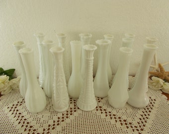 May include: A collection of 12 white milk glass bud vases, arranged on a crocheted doily. The vases are all different shapes and sizes, and some have a textured design.