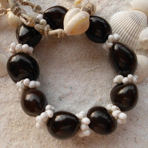 May include: A dark brown seed bracelet with white shell beads. The bracelet is on a white sandy surface.