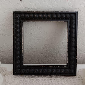 May include: A square picture frame with a black finish and a decorative beaded border. The frame features visible distressing along the edges, revealing lighter tones beneath the dark paint, creating a textured appearance around the central opening.
