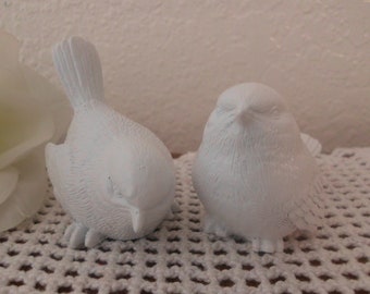 May include: Two white ceramic bird figurines, one with its head tilted down and the other with its head tilted up, sitting on a white crocheted surface.