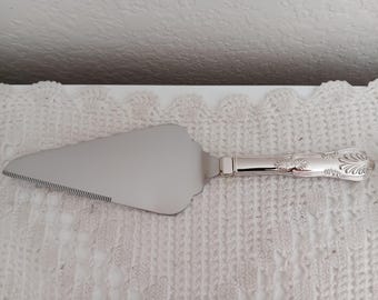 May include: Silver cake server with a serrated edge and ornate handle. The server has a wide, triangular blade and a decorative floral pattern on the handle. The server is displayed on a white, textured surface.