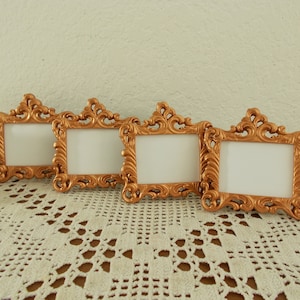 May include: Four gold-colored ornate picture frames with white mats. The frames are arranged in a row on a white crocheted doily.