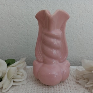 May include: A glossy, light pink ceramic vase featuring a central twisted, rope-like column design. The vase has a wide, bulbous base and a flared, scalloped rim at the top. The surface is smooth and reflective, showcasing a solid pastel pink color.