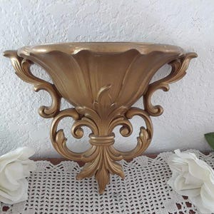 May include: A gold-colored wall-mounted planter with a decorative, ornate design. The planter has a scalloped rim and a fluted bowl.