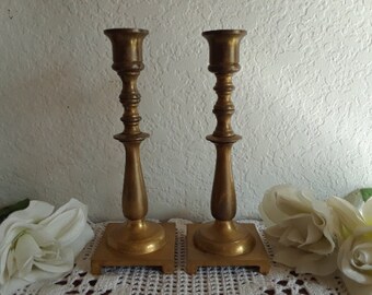 May include: Two gold metal candle holders with a simple design. The candle holders have a round base and a tall, slender stem.