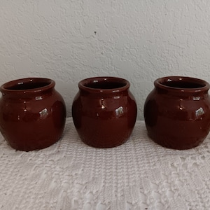 May include: Three small, brown ceramic pots with a glossy finish. The pots are arranged in a row on a white crocheted surface.