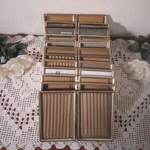 May include: A collection of small, rectangular vintage display boxes with gold-colored frames. The boxes have various interior designs, including ribbed paper and plain white inserts. The arrangement suggests a display or storage solution.