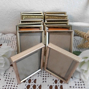 May include: A collection of small, rectangular gold-toned picture frames. Some frames are stacked, while others are arranged in front. The frames have a simple, elegant design, with a thin border and a glass or plastic insert. The background includes white flowers and a starfish.