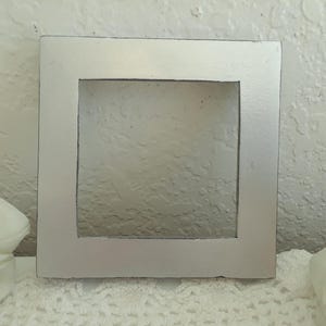 May include: A silver picture frame with a square opening. The frame is made of wood and has a smooth, painted finish.