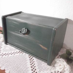 May include: A dark green wooden bread box with a leaf-shaped metal handle. The box is sitting on a white crocheted doily.