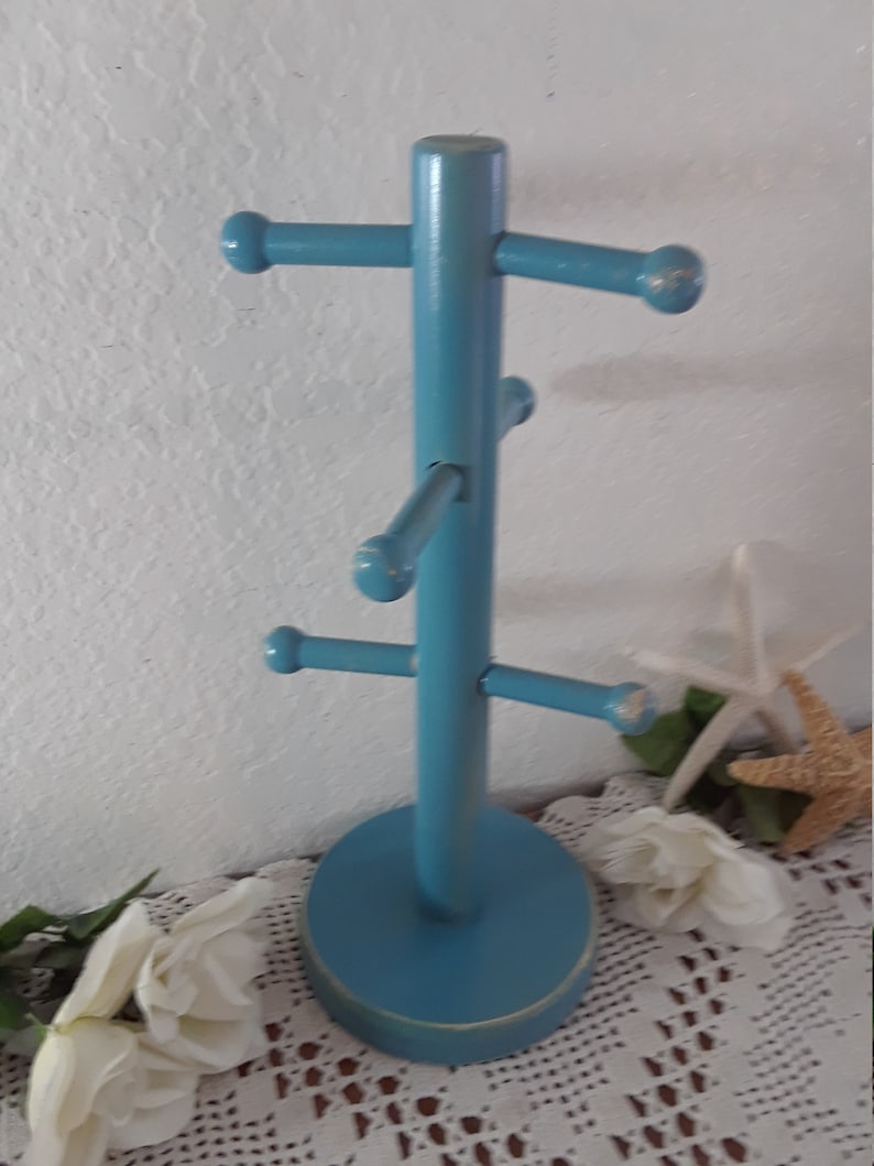 May include: A blue wooden mug tree with five hooks for hanging mugs. The tree has a round base and is painted a light blue color.