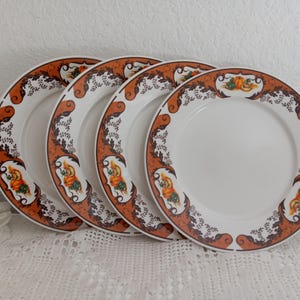 May include: Four white ceramic plates with a decorative orange and brown border. The border features a repeating pattern of figures and foliage. The plates are arranged on a white lace tablecloth.