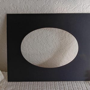 May include: A black picture frame with an oval opening, revealing a textured white surface. The frame is rectangular and sits on a white lace tablecloth. The background is a white wall.