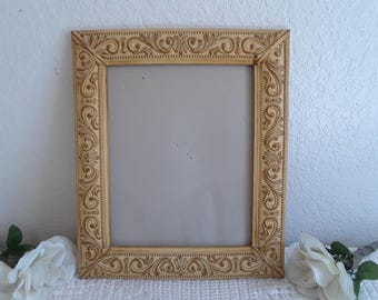 May include: A decorative picture frame with an ornate, carved design. The frame is painted in a light beige color and has a rectangular shape.