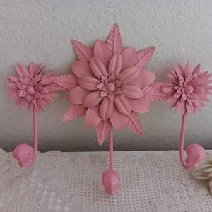 May include: A pink metal wall hook with a floral design. The hook features three flower-shaped elements, with a central, larger flower and two smaller ones on either side. Each flower has a hook below for hanging items.