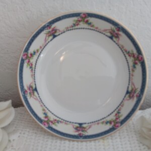 May include: A white ceramic bowl featuring a wide rim decorated with a dark blue border, gold trim, and a repeating pattern of pink roses, green leaves, and light blue ribbons. The center of the bowl is plain white with a smooth, glossy finish.