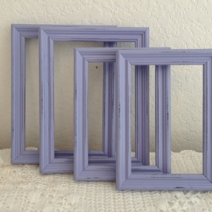 May include: Four lavender-colored picture frames of varying sizes, with a distressed finish, arranged in a row on a white lace tablecloth.