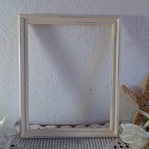 May include: A white, distressed wood picture frame with a simple, rectangular design. The frame is sitting on a white crocheted surface with white flowers and starfish.