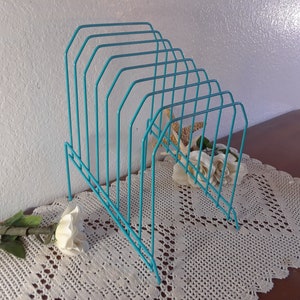 May include: A turquoise blue metal magazine rack with eight slots for holding magazines or papers. The rack is on a white crocheted doily on a wooden surface.