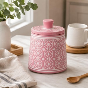 Può includere: Un barattolo cilindrico in ceramica rosa con coperchio abbinato, caratterizzato da un motivo geometrico bianco in rilievo composto da cerchi intrecciati e piccole forme a stella. Il barattolo ha una finitura liscia e un pomello centrale.