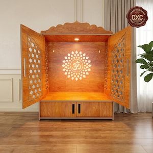 Könnte beinhalten: Ein hölzerner Tempelschrank mit zwei offenen Türen, die komplizierte Gitterausschnitte aufweisen. Die Rückwand zeigt ein leuchtendes Om-Symbol, umgeben von einem Sonnenstrahlenmuster. Ein kleines Licht ist oben integriert und der Sockel enthält zwei Aufbewahrungsschubladen mit vertikalen schwarzen Griffen.