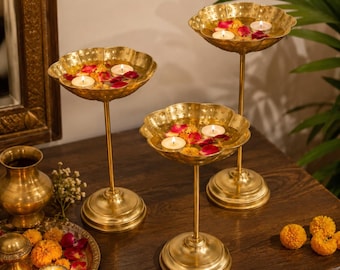 Puede incluir: Tres cuencos de metal dorado sobre pedestal con bordes festoneados, cada uno con agua, velas de té flotantes y pétalos de flores rojos y amarillos. También se incluyen un jarrón dorado a juego y una pequeña bandeja con flores.