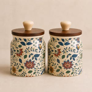 May include: Two cylindrical ceramic canisters featuring a cream-coloured base with a repeating pattern of blue, red, and green floral vines and leaves. Each canister is topped with a dark brown wooden lid and a small, light-coloured wooden knob.