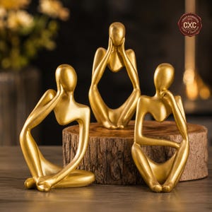 Pode incluir: Três esculturas douradas de figuras humanas abstratas em poses sentadas. Uma figura cobre os ouvidos, outra os olhos e a terceira apoia o queixo na mão. As esculturas são exibidas em uma superfície de madeira.