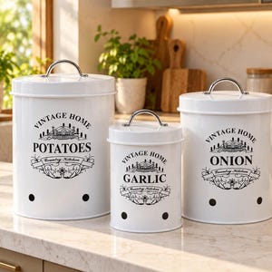 Könnte beinhalten: Ein Set aus drei weißen, zylindrischen Vorratsbehältern aus Metall mit silbernen, bogenförmigen Griffen auf den Deckeln. Jeder Behälter trägt den schwarzen Schriftzug Vintage Home, Country Kitchen sowie die Aufschriften Potatoes, Garlic und Onion.