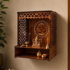 Könnte beinhalten: Ein hängender Holzschrein mit aufwendigen Gitterseitenwänden und einem Sonnenstrahlen-Design mit Om-Symbol auf der Rückwand. Im Inneren befinden sich eine Ganesha-Statue aus Messing, eine kleine Glocke, Räucherstäbchen und zwei kleine Messingschalen.