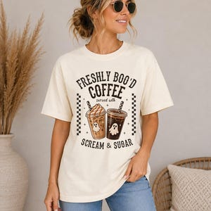 May include: A cream-colored short-sleeve t-shirt featuring a graphic print with the text "FRESHLY BOO'D COFFEE served with SCREAM & SUGAR". The design includes two iced coffee cups with ghost illustrations, surrounded by small stars and checkered borders.