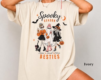 May include: An ivory t-shirt featuring a graphic print with the text "Spooky Season" and "Emma Sarah Olivia BESTIES". The design shows three skeleton figures dressed in Halloween costumes, including a ghost and a witch, with a black cat.