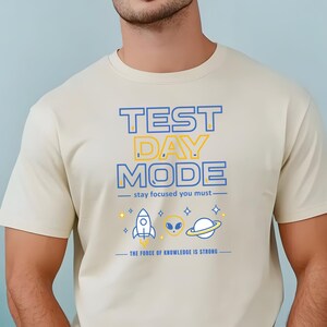 May include: Beige t-shirt with the text "TEST DAY MODE" in blue and yellow. The shirt features a rocket ship, alien, and planet graphic. The phrase "stay focused you must" and "THE FORCE OF KNOWLEDGE IS STRONG" are also printed on the shirt.
