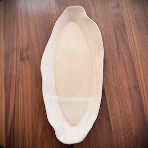 May include: Off-white, oblong ceramic serving dish with a textured surface. The dish has a raised rim and a unique, organic shape. The dish is placed on a dark brown wooden table.