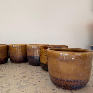 May include: A collection of handmade ceramic cups with a glossy, glazed finish. The cups are a warm brown color with a darker, purplish-brown band near the base. The cups are arranged in a row on a speckled surface.