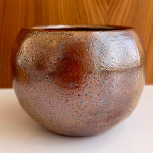 May include: A round ceramic bowl with a glossy, mottled finish in shades of copper, bronze, and dark brown. The surface features a textured, speckled appearance with subtle variations in tone, creating a reflective, metallic-like sheen across the exterior.