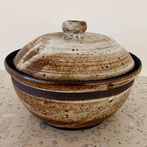 May include: A ceramic serving bowl with a lid. The bowl and lid are primarily beige with brown accents and speckles. The bowl has a dark brown band around the middle. The lid has a small, rounded handle.