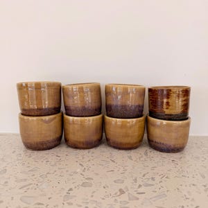 May include: A collection of eight small, handmade ceramic bowls. The bowls are a warm, earthy brown with a darker, purplish-brown band near the base. The glaze gives them a glossy finish, and they are stacked in two rows.