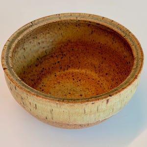May include: A handmade ceramic bowl with a speckled, earthy design. The bowl features a light green exterior with a brown interior and rim. The bowl is a perfect size for serving snacks or small dishes.