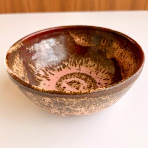 May include: A handmade ceramic bowl with a unique glaze. The bowl features a reddish-brown rim that transitions to a speckled brown and tan exterior. The interior has a pinkish-tan base with a darker brown spiral design. This decorative bowl is perfect for serving or display.