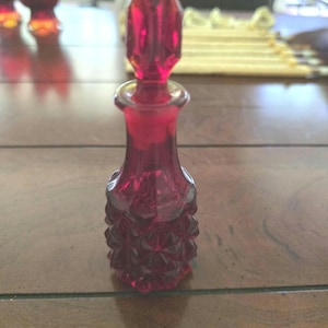 Op de afbeelding: Een klein parfumflesje van dieprood glas met een gefacetteerd, diamantachtig patroon op het onderste gedeelte en een bijpassende puntige, geometrische glazen stop. Het flesje heeft een slanke hals en een gladde, uitlopende rand, met een levendige, doorschijnende robijnrode kleur over het gehele object.
