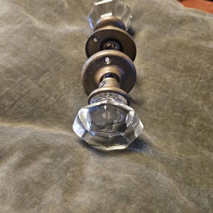 May include: A door knob set featuring two clear, multifaceted glass knobs with an octagonal shape. The knobs are connected by a central metal spindle and include two circular, aged brass-toned mounting plates with visible screw holes for installation.